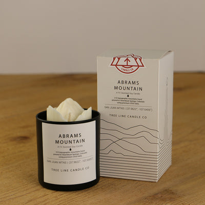 A white wax candle named Abrams Mountain is next to a white box with red and black lettering.