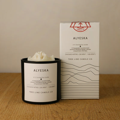 A white wax candle named Alyeska is next to a white box with red and black lettering.