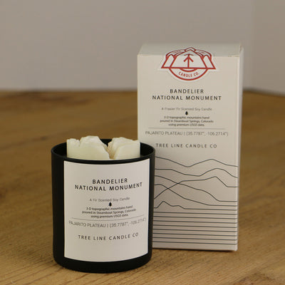 A white wax candle named Bandelier National Monument is next to a white box with red and black lettering.
