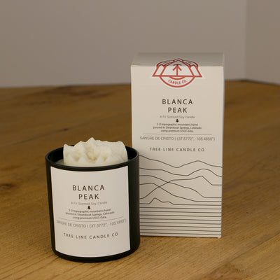 A white wax candle named Blanca Peak is next to a white box with red and black lettering