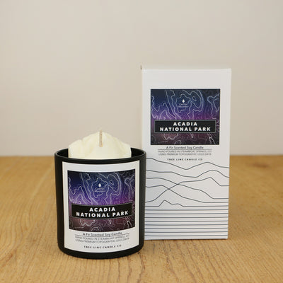 A white wax candle of Cadillac Mountain with a purple and black label is next to a white box with topography lines named Acadia National Park by Tree Line Candle Co.