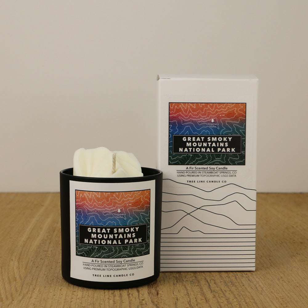 A white wax candle of Clingmans Dome with an orange, blue and green label is next to a white box with topography lines named Great Smokey Mountains National Park by Tree Line Candle Co.