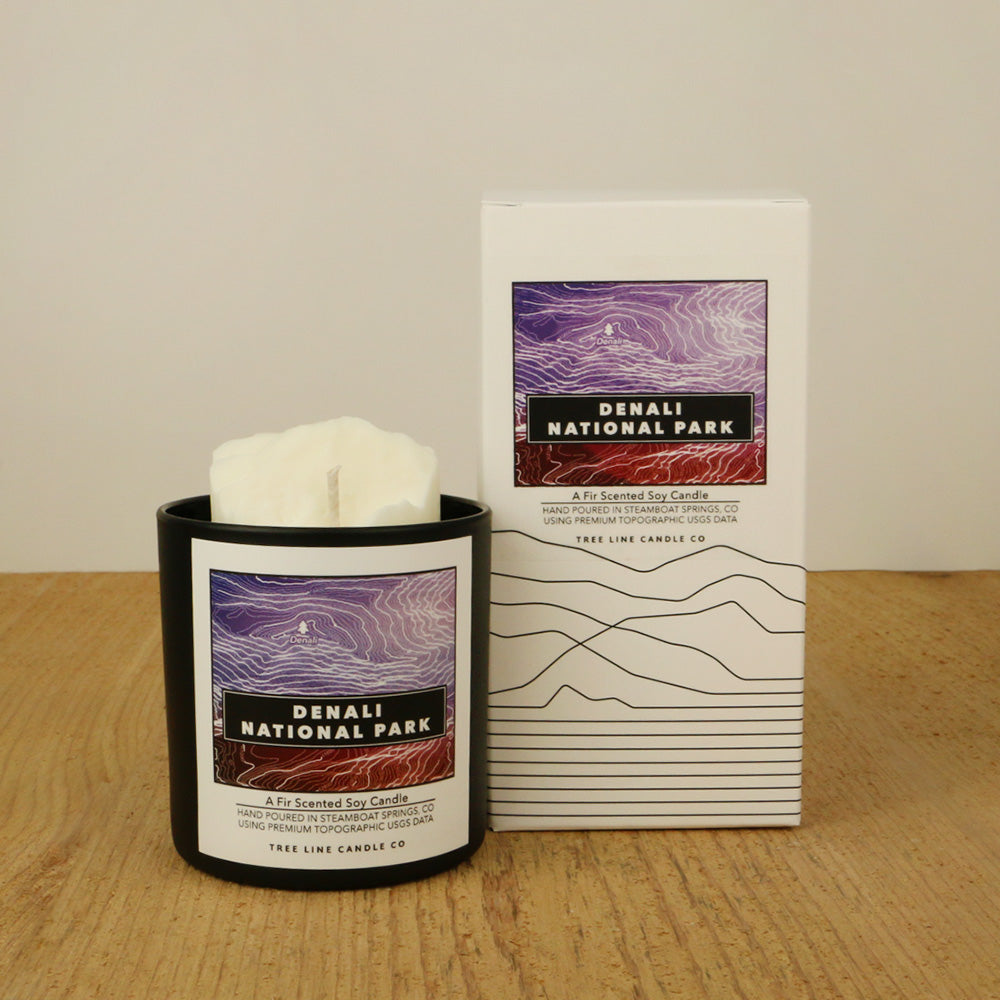 A white wax candle of Denali with a purple and maroon label is next to a white box with topography lines named Denali National Park by Tree Line Candle Co.