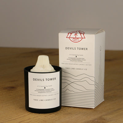 A white wax candle named Devils Tower is next to a white box with red and black lettering.
