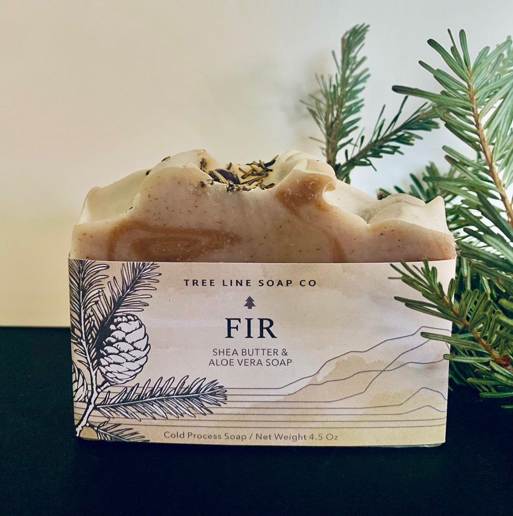 A single bar of Fir soap is next to a fir sprig on a daark blue cloth.