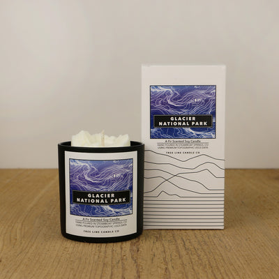 A white wax candle of Mount Grinnell with a purple and black label is next to a white box with topography lines named Glacier National Park by Tree Line Candle Co.