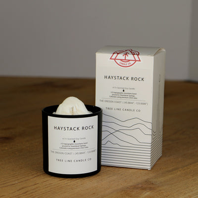 A white wax candle named Haystack Rock is next to a white box with red and black lettering.