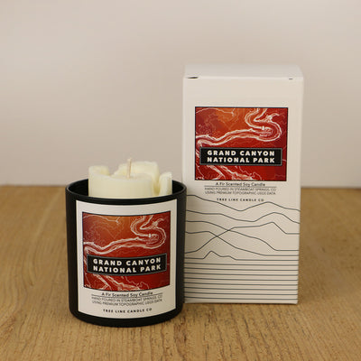 A white wax candle of Horseshoe Bend with an orange and red label is next to a white box with topography lines named Grand Canyon National Park by Tree Line Candle Co.