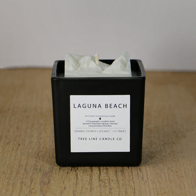 A white candle replica of Laguna Beach cliffs and shore in a square, black container.