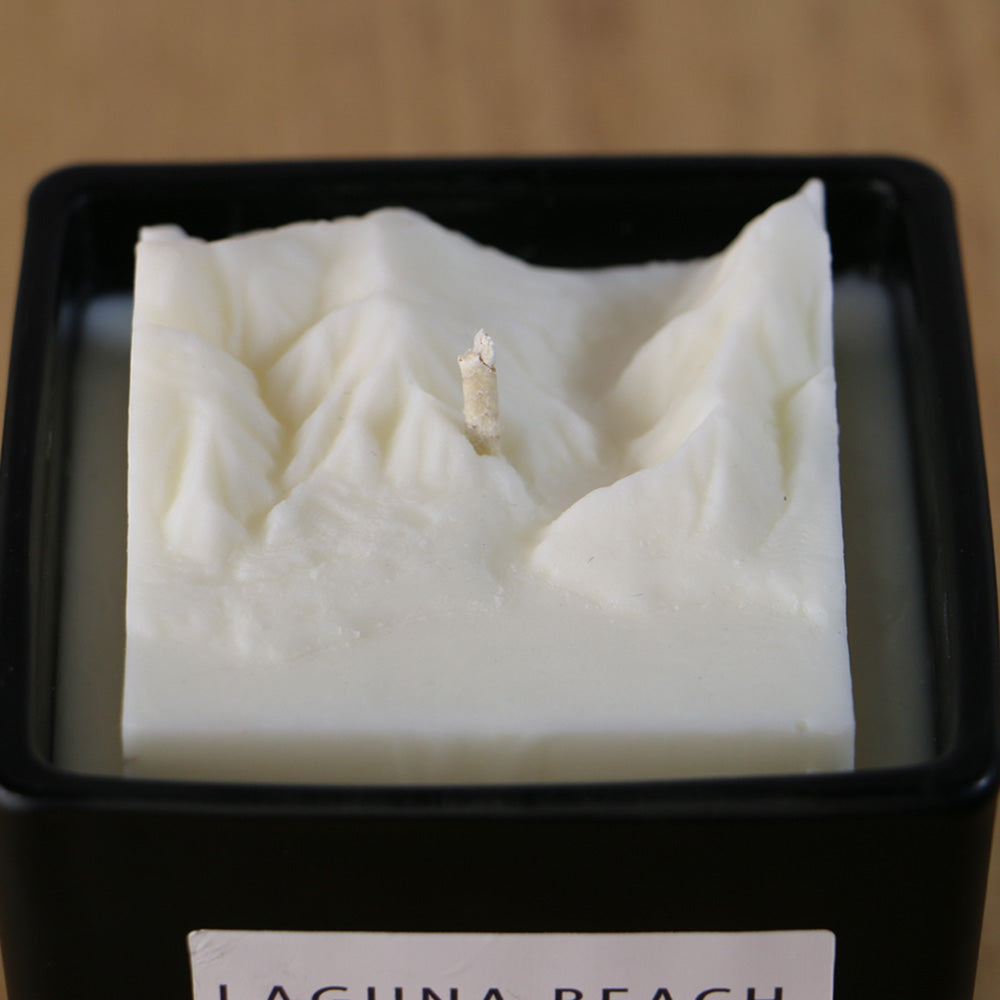 A close-up view of the white candle replica of Laguna Beach cliffs and shore in a square, black container.