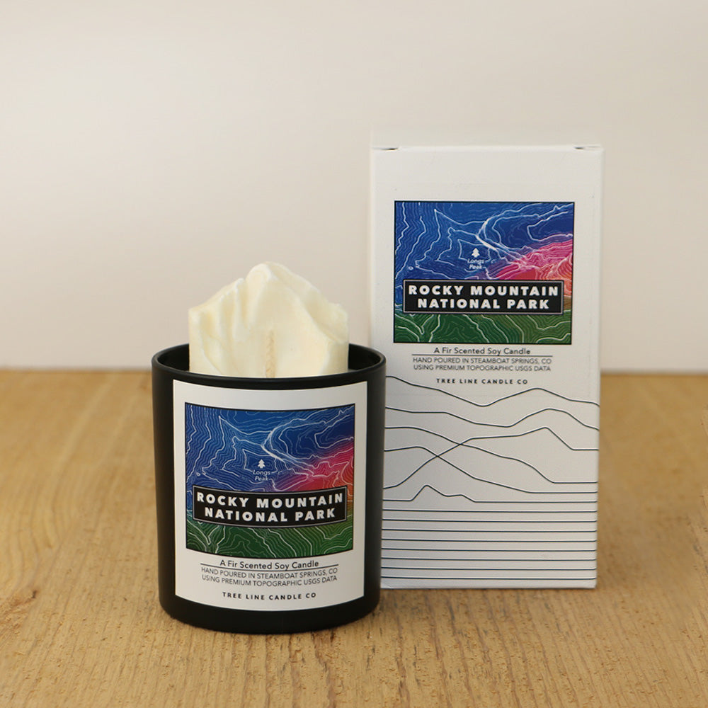 A white wax candle of Longs Peak with a blue, red-orange and green label is next to a white box with topography lines named Rocky Mountain National Park by Tree Line Candle Co.