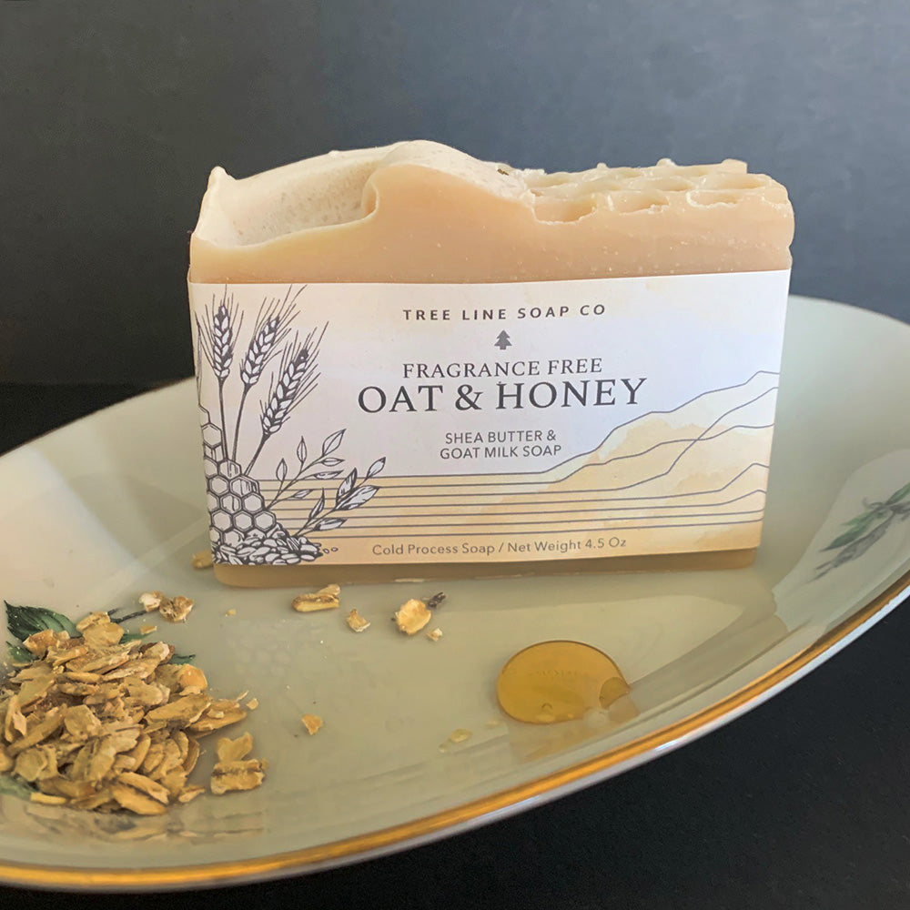 A bar of Oat & Honey soap stands behind a bit of oats and  a drop of honey on a china dish.