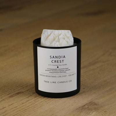 A white soy wax replica candle of Sandia Crest in a round, black glass.