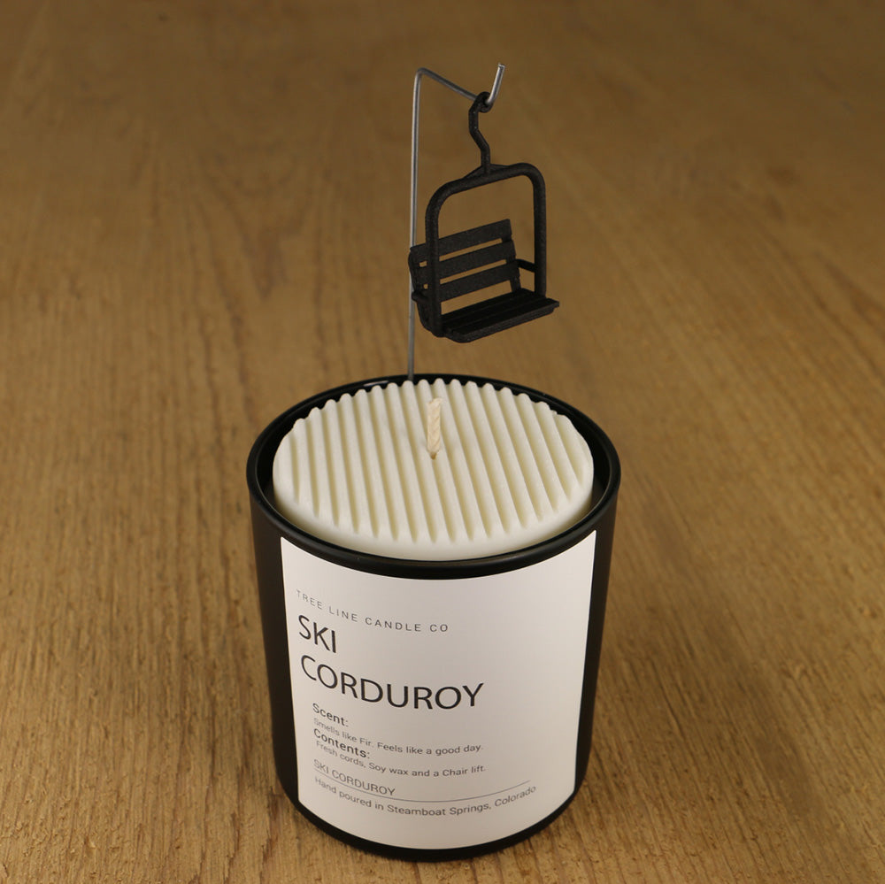 A white soy wax replica candle of SKI Corduroy in a round, black glass.