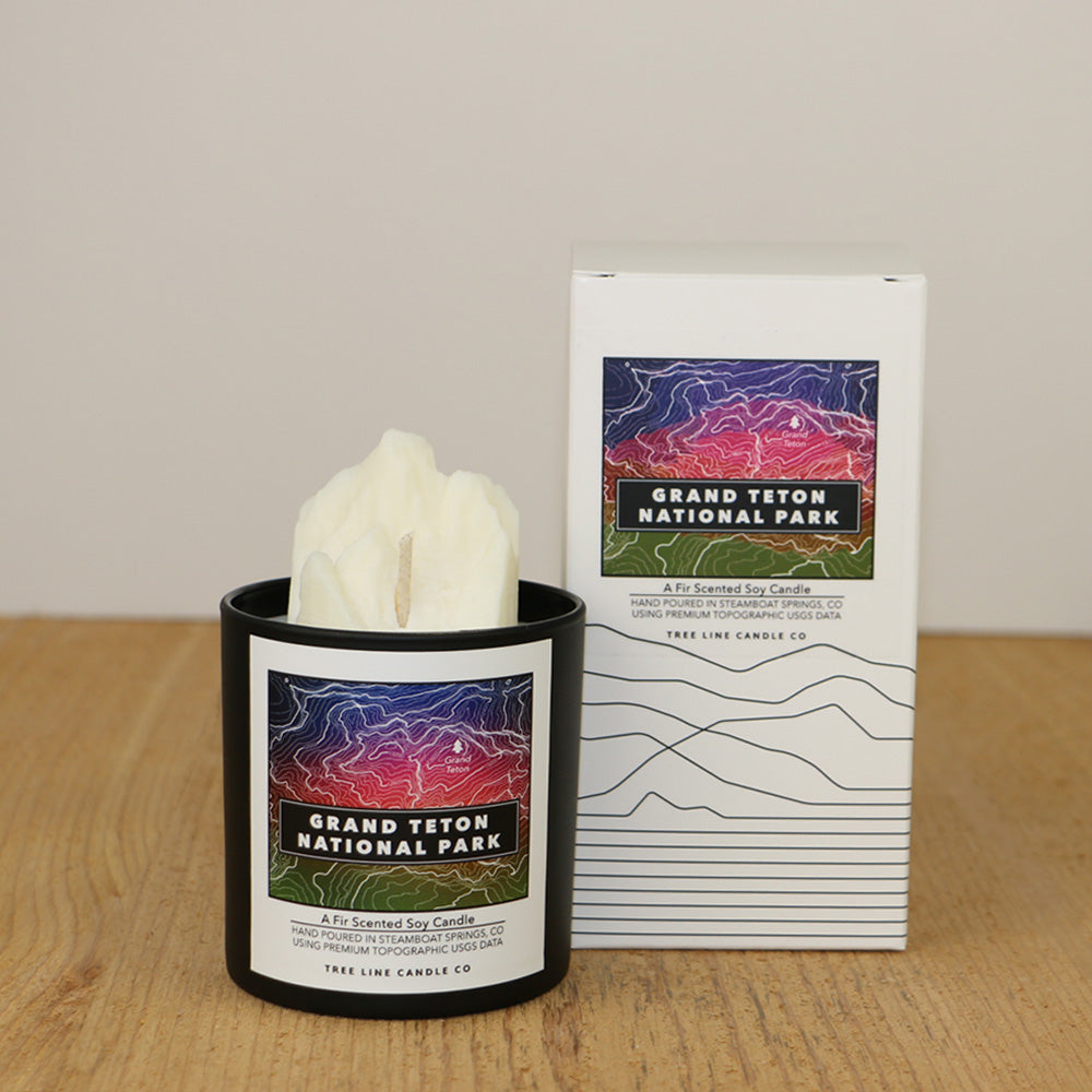 A white wax candle of Grand Teton with a purple, red and green label is next to a white box with topography lines named Grand Teton National Park by Tree Line Candle Co.